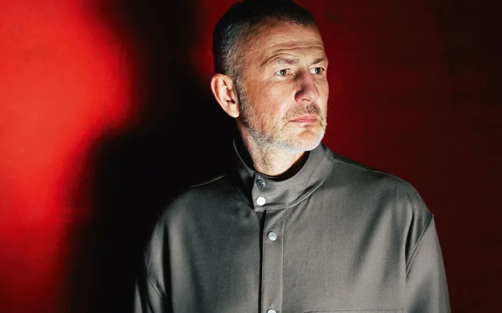A man with a shaved head stands against a deep red background, lit dramatically.