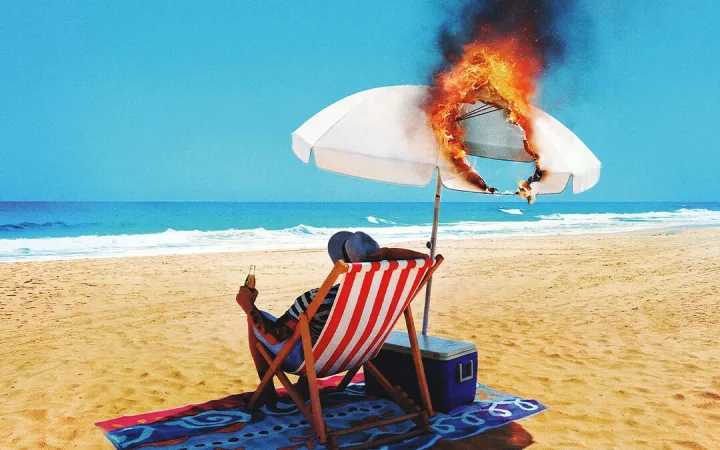 Person relaxing in a deckchair on a beach as the parasol above burns fiercely.