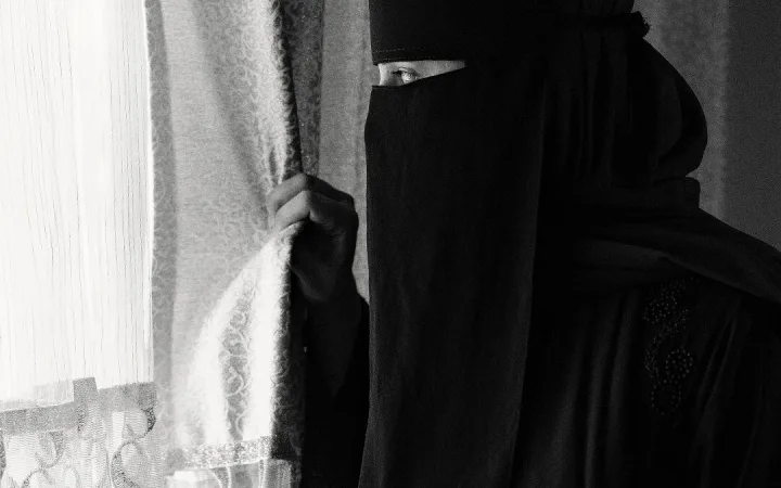 A woman wearing a niqab stands near a window, photographed in black and white.