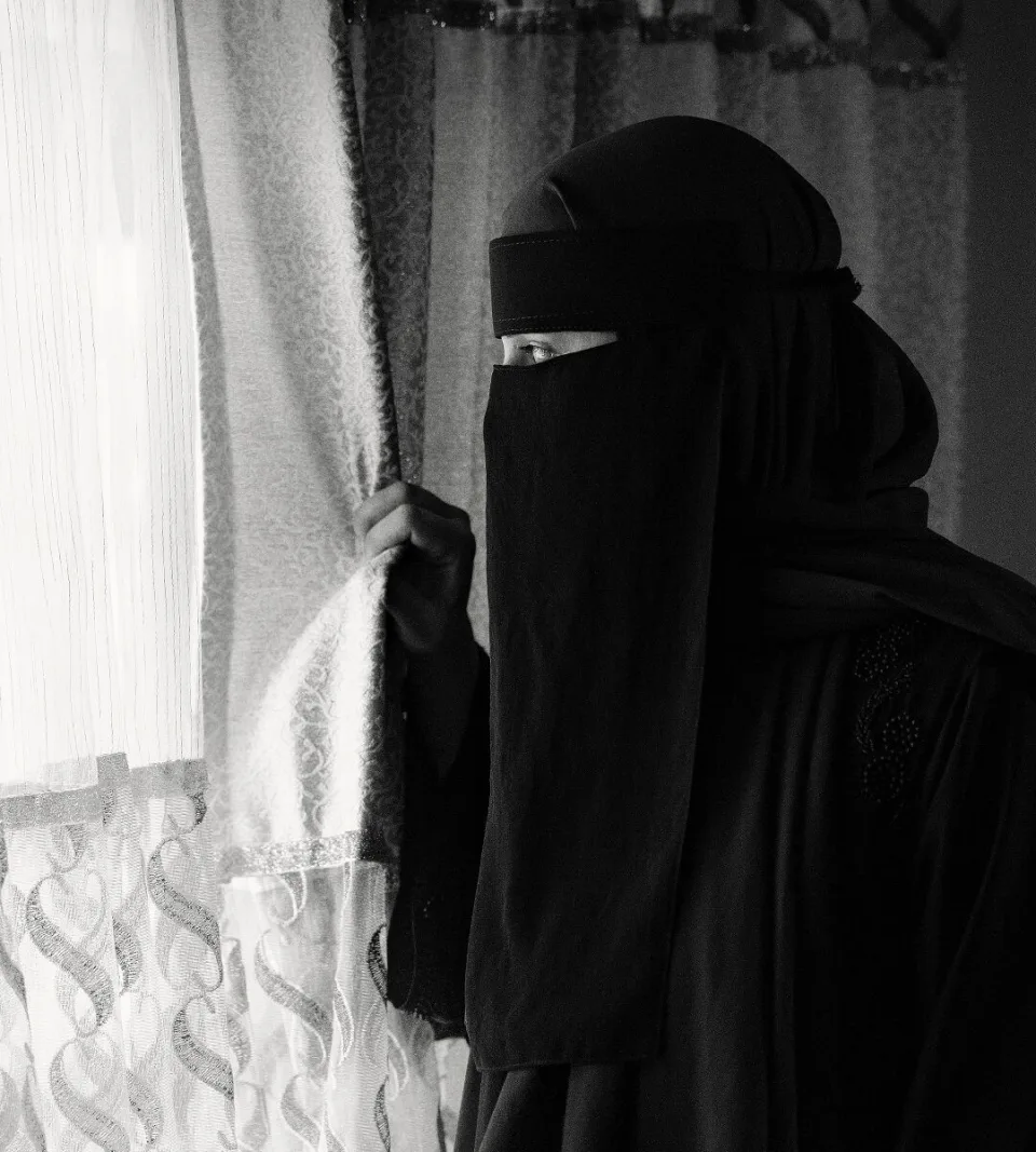 A woman wearing a niqab stands near a window, photographed in black and white.