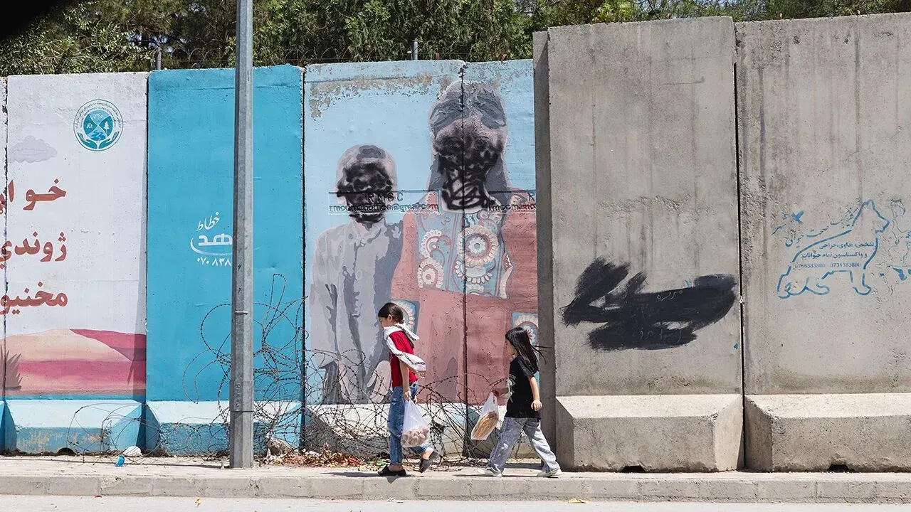 Two girls walk along a road in Kabul past a mural in which the human faces have been obscured with black paint