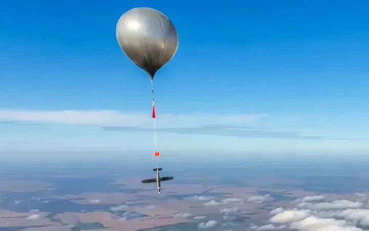 Silvery high-altitude balloon drifting above clouds in clear blue sky.